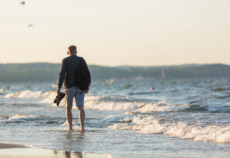 Man walking along a shore holding his shoes.